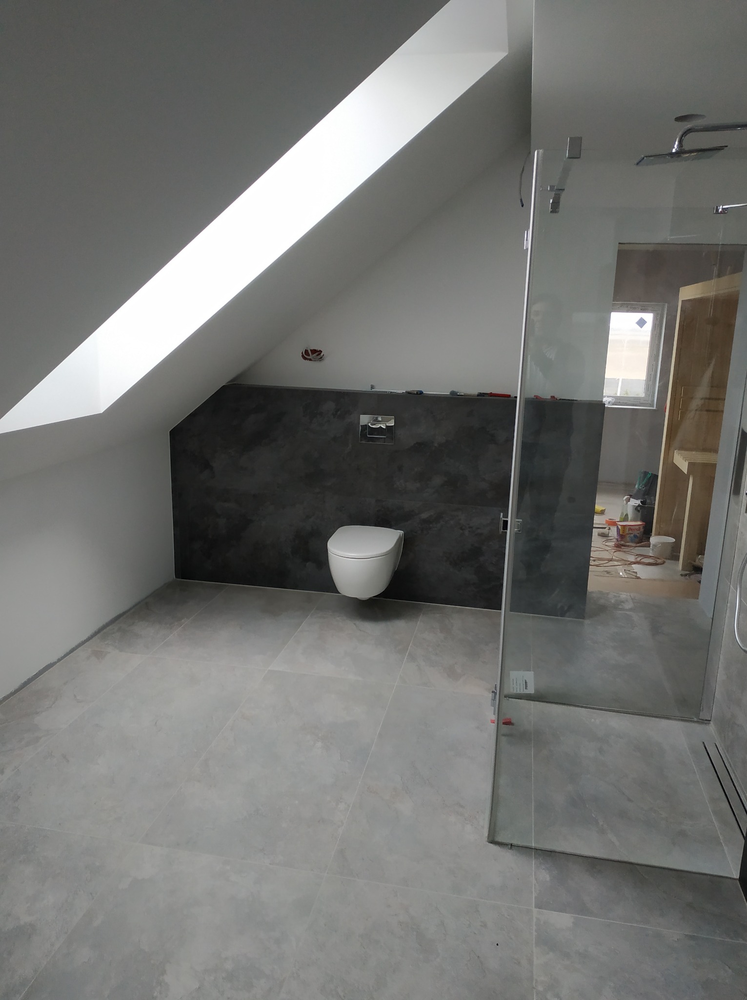 Nowoczesna łazienka na poddaszu z szarą płytką podłogową, ścianą w ciemnym tynku dekoracyjnym, kabiną prysznicową ze szkła i wiszącą toaletą. Widoczne narzędzia i materiały budowlane w tle.