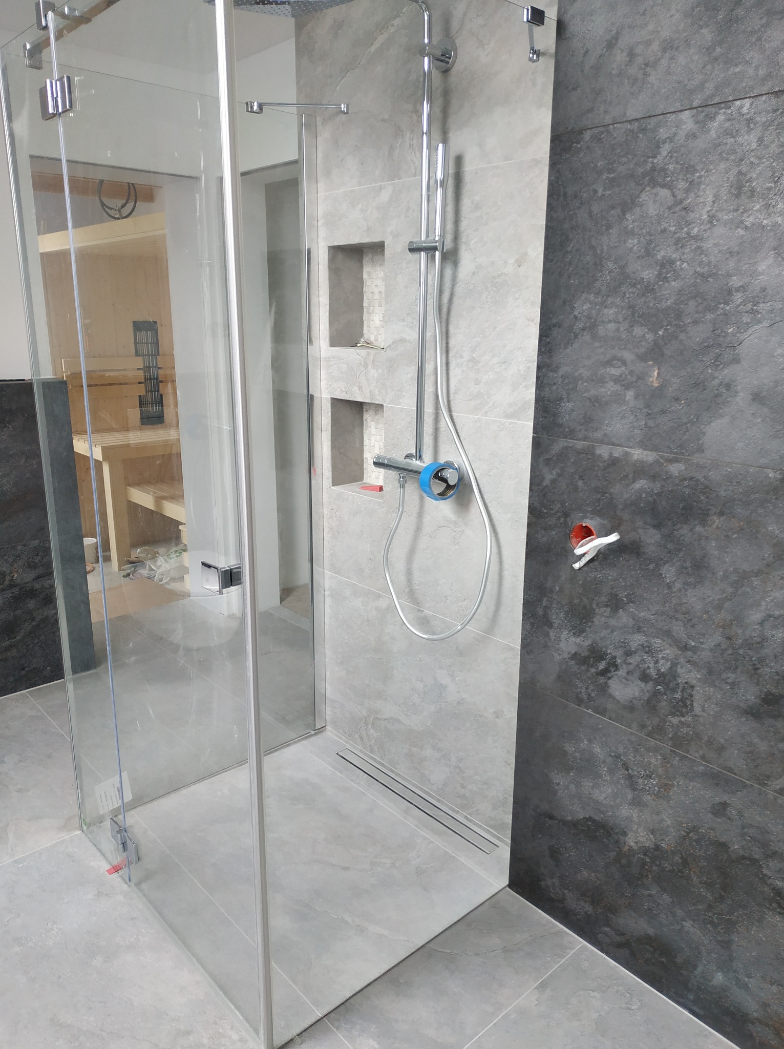 Nowoczesna kabina prysznicowa z odpływem liniowym, szare płytki imitujące kamień, widoczna instalacja prysznicowa z niebieskim zabezpieczeniem na baterii, w tle fragment sauny w trakcie budowy.