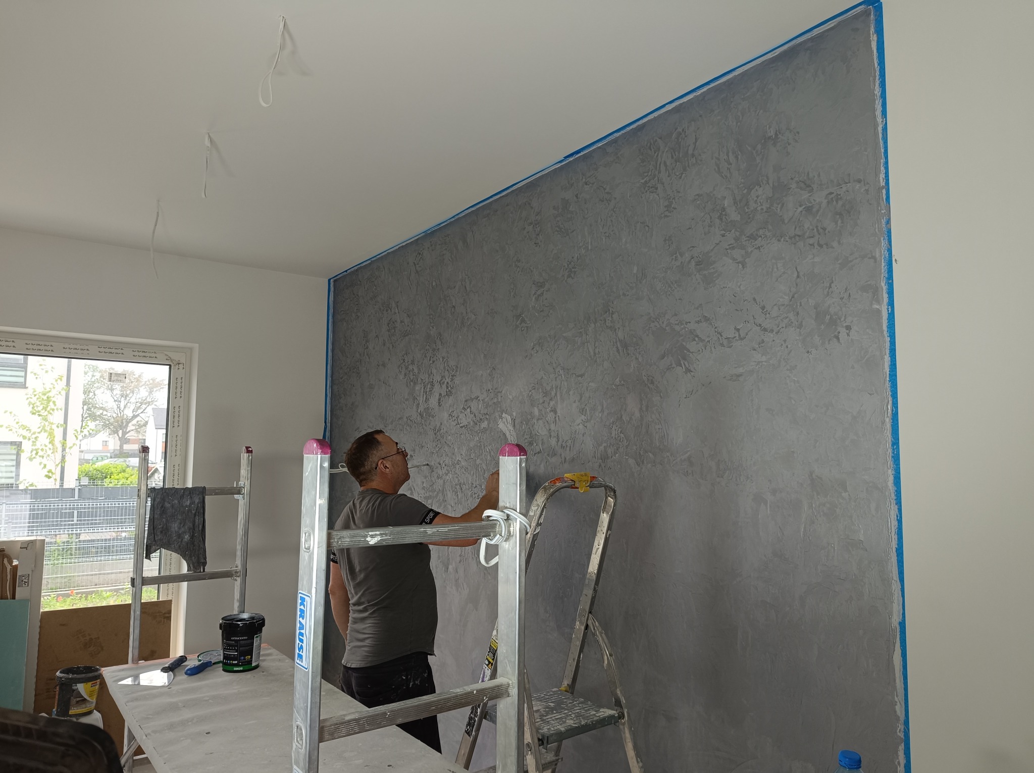 Pracownik nanosi dekoracyjną, szarą strukturę na ścianę, zabezpieczoną niebieską taśmą malarską, używając szpachelki; widoczne drabiny, stół roboczy z narzędziami i puszkami farby.