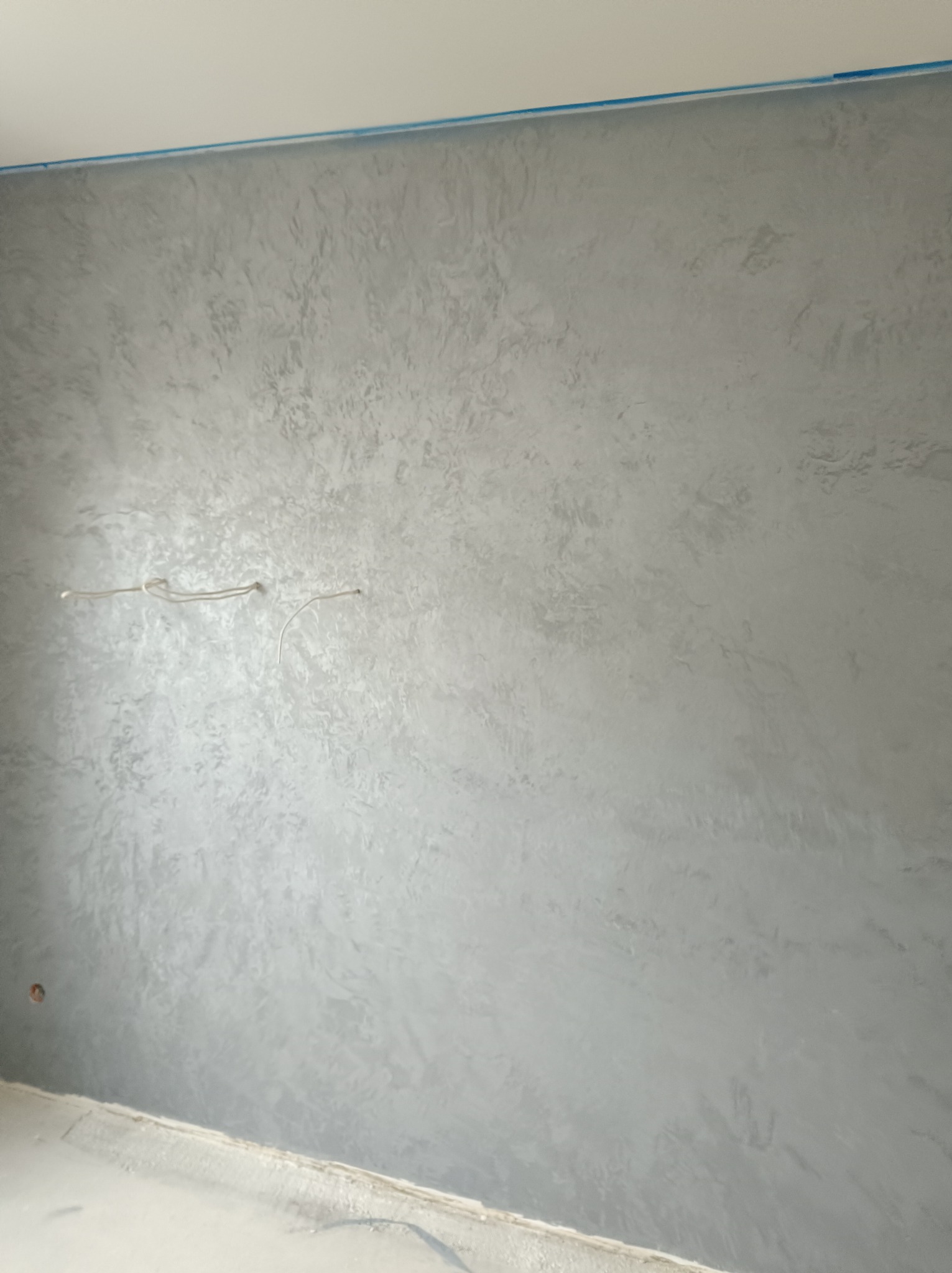 Ściana pokryta dekoracyjnym tynkiem w odcieniach szarości, z widocznym przewodem elektrycznym i zabezpieczeniem taśmą malarską przy suficie.