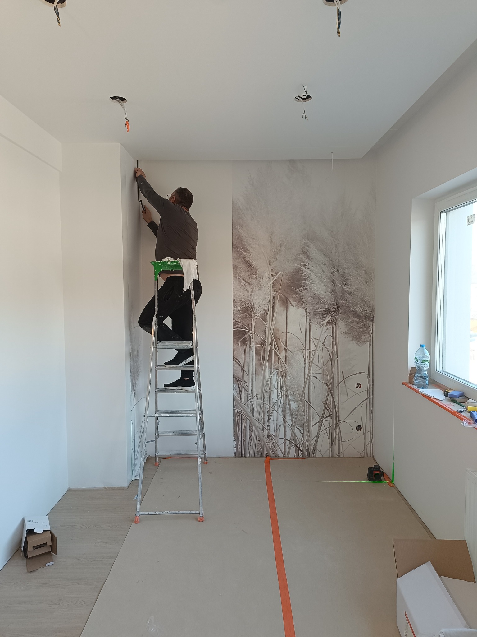 Pracownik na drabinie przykłada tapetę z motywem traw do ściany, widoczny laser krzyżowy wyznaczający linię, surowy stan wykończenia, niepodłączone kable oświetleniowe.