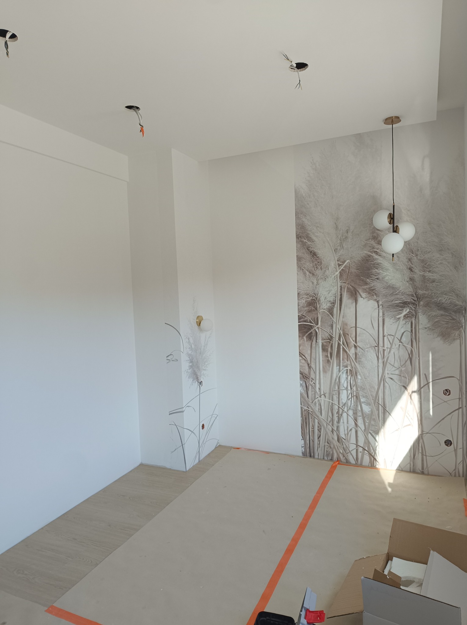 Pokój w trakcie wykańczania z położoną tapetą w motywy roślinne, widoczne kable oświetleniowe wystające z sufitu, podłoga zabezpieczona kartonem, ściany pomalowane na biało.