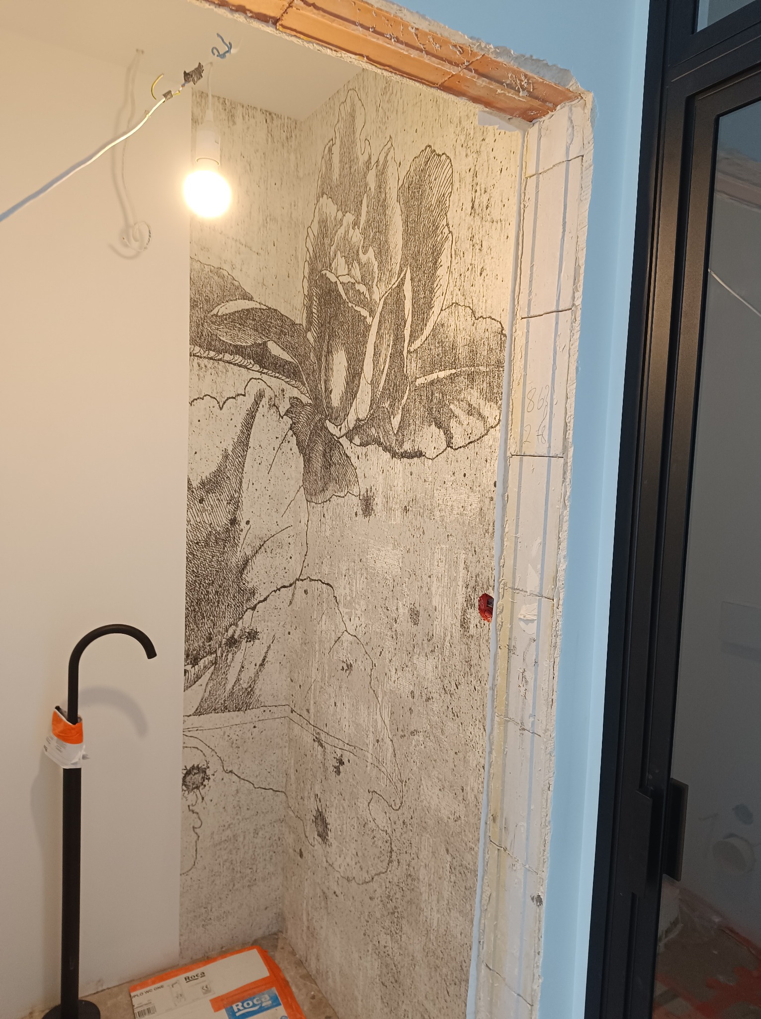 Przygotowana ściana w łazience z dekoracyjnym rysunkiem kwiatowym przed położeniem płytek, widoczna rura odpływowa Roca i surowy otwór drzwiowy z niebieskim obramowaniem.