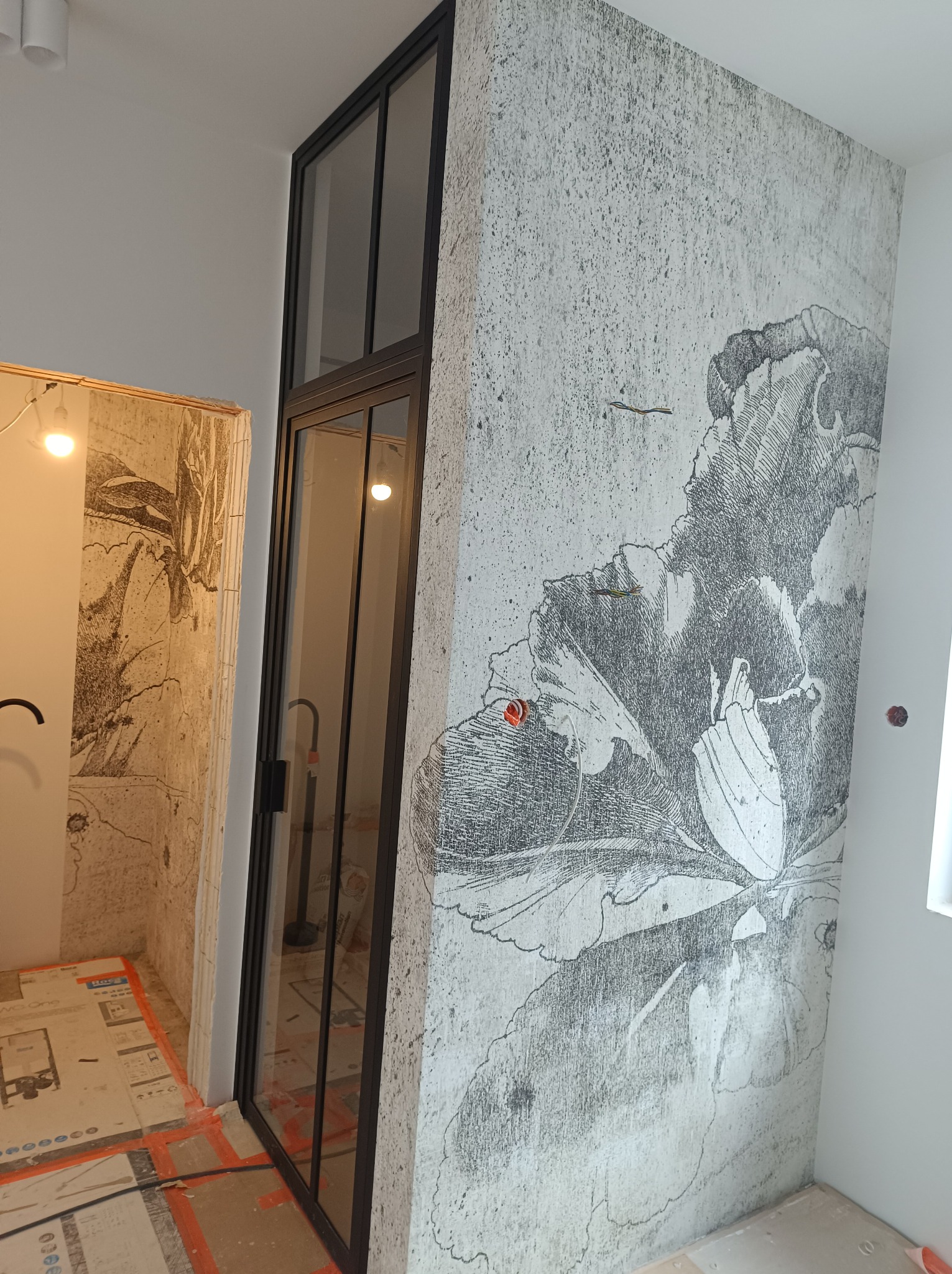 Ściana z szarą tapetą w motywy kwiatowe i czarna, przeszklona rama drzwi w trakcie prac wykończeniowych, widoczne kable i zabezpieczenia podłogi.