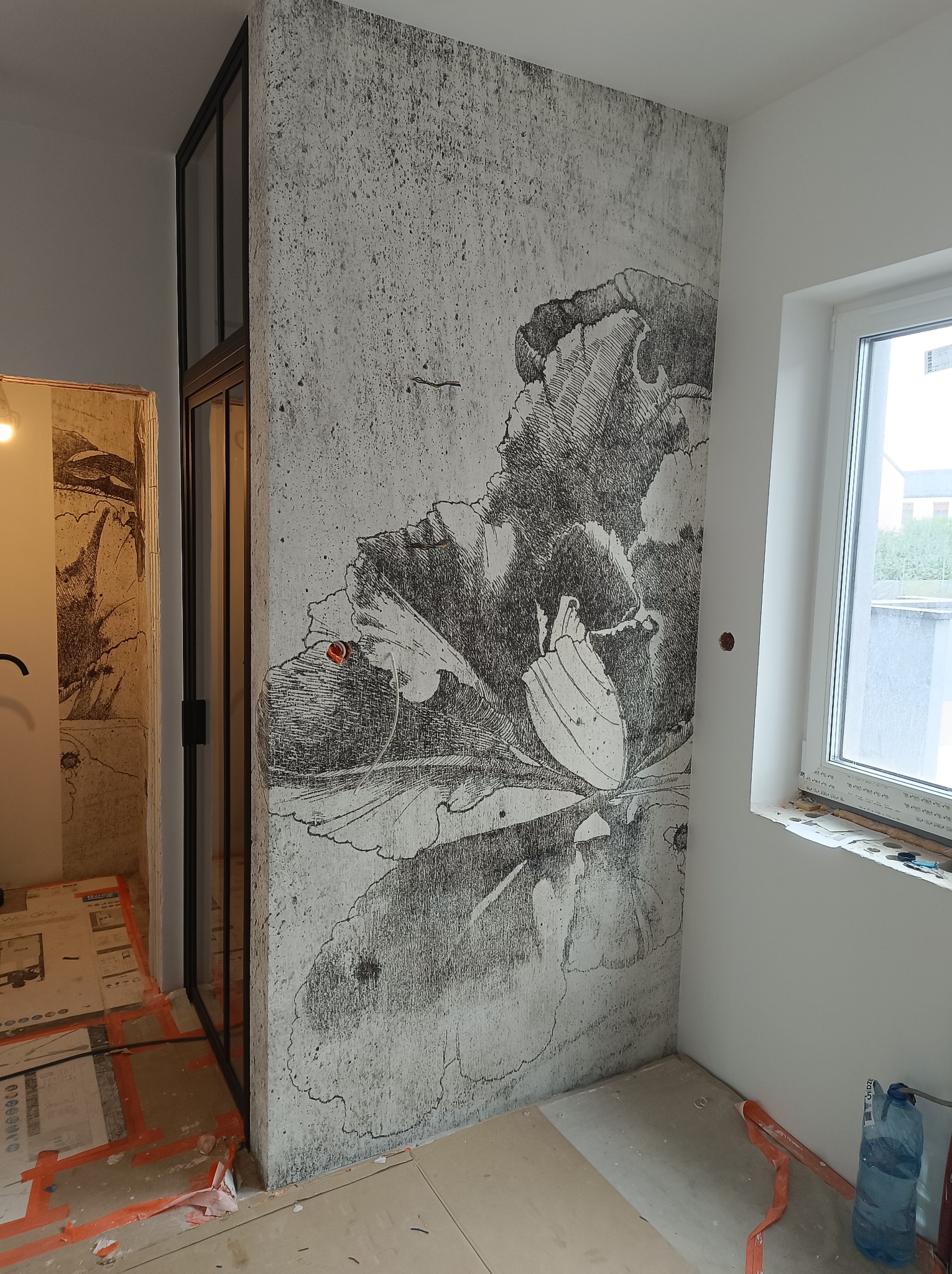 Fragment ściany z fototapetą o motywie kwiatowym w stylu szkicu, obok okno i przeszklona konstrukcja w trakcie prac wykończeniowych, widoczne zabezpieczenia podłogi.