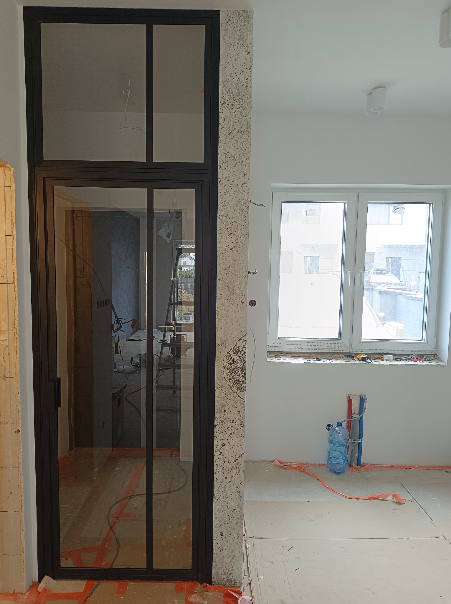 Montaż szklanej ścianki działowej z czarnymi profilami we Wrocławiu, widoczne przygotowania do dalszych prac wykończeniowych, zabezpieczona podłoga, instalacja wodna bez armatury.