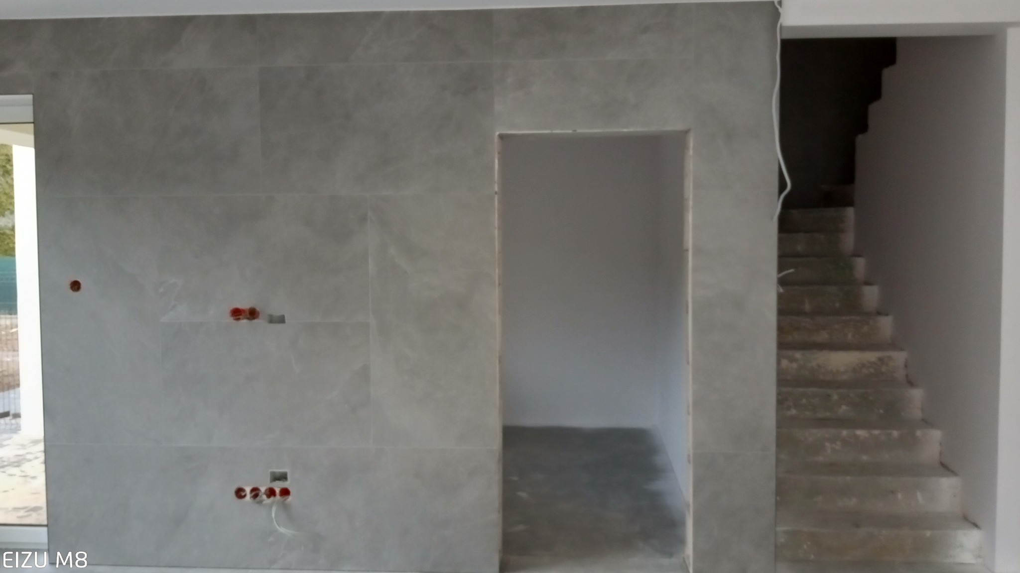 Ściana wyłożona szarymi płytkami imitującymi kamień z przygotowanymi otworami na gniazdka elektryczne i wnęką drzwiową, widok na betonowe schody w głębi pomieszczenia.