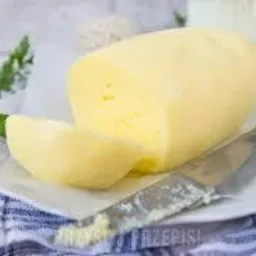 Blok żółtego sera z odciętym plastrem, leżący na białym talerzu z niebieską kratą, obok nóż i gałązka zielonej pietruszki, w tle mleko.