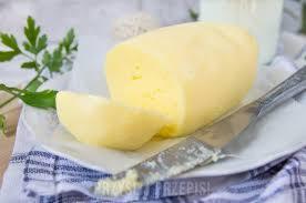 Blok żółtego sera z odciętym plastrem, leżący na białym talerzu z niebieską kratą, obok nóż i gałązka zielonej pietruszki, w tle mleko.