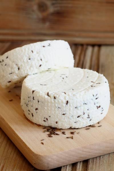 Dwa okrągłe sery twarogowe z kminkiem na drewnianej desce. Jeden ser częściowo zasłonięty, drugi w całości widoczny. Tło z drewnianych desek.