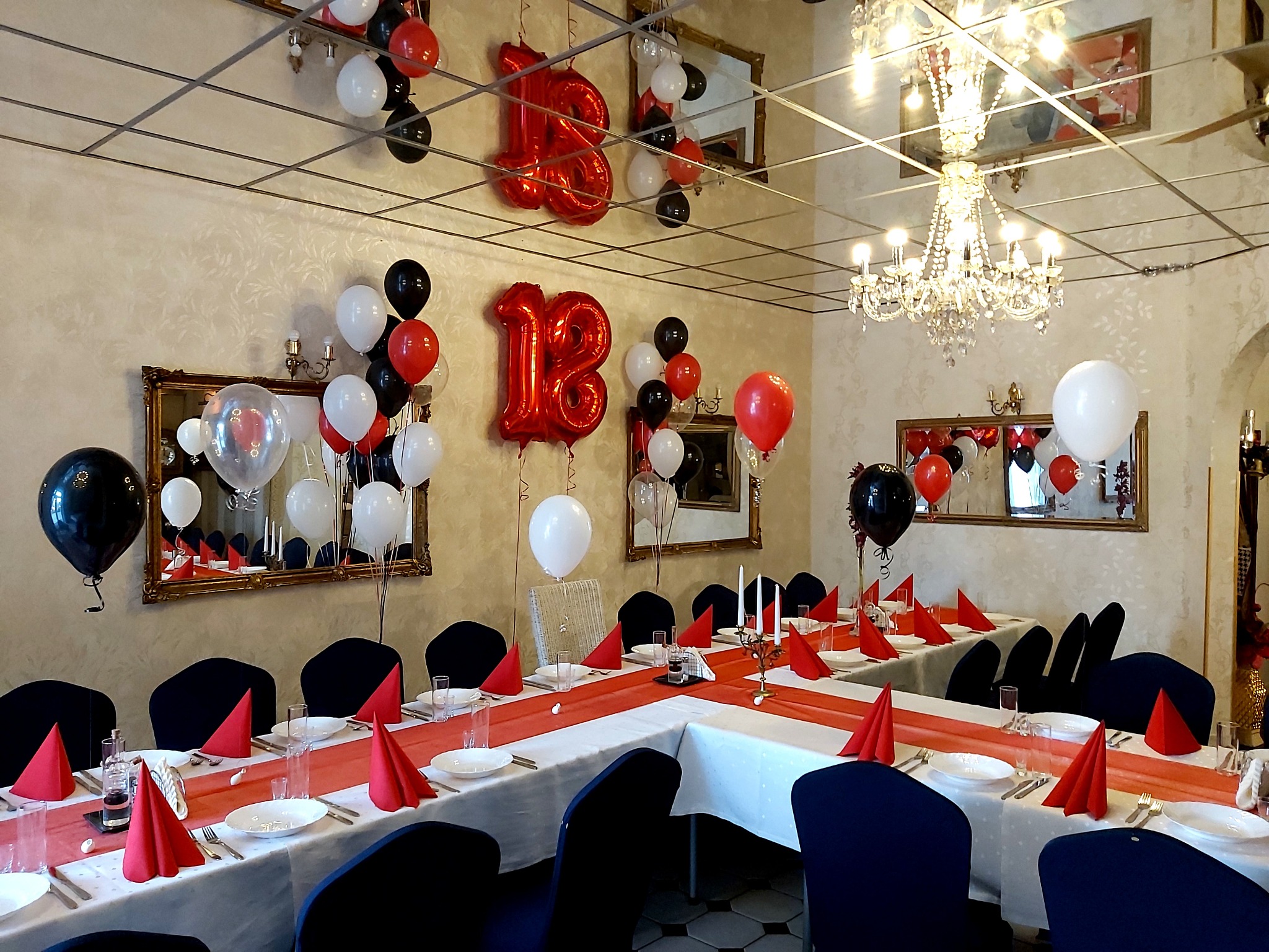 Sala udekorowana na osiemnaste urodziny: czerwone balony w kształcie cyfr '18', białe, czerwone i czarne balony, elegancko nakryty stół z czerwonymi serwetkami i bieżnikiem, lustrzany sufit...