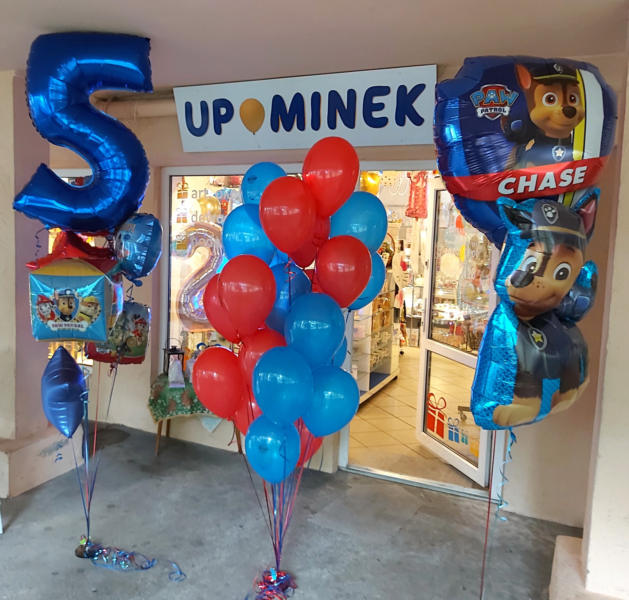 Dekoracja balonowa przed wejściem do sklepu z upominkami, z balonami w kształcie cyfry 5, balonami z motywem Psi Patrol i bukietem czerwono-niebieskich balonów.