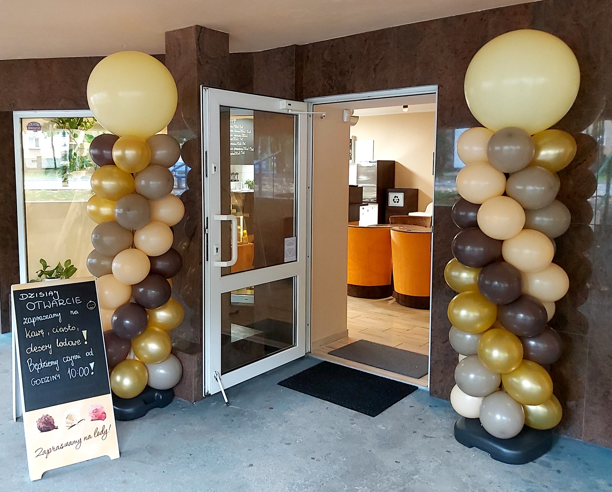 Dekoracja balonowa w odcieniach złota, brązu i beżu przed wejściem do lokalu w Szczecinie, zapraszająca na otwarcie i lody, z tablicą informacyjną obok.