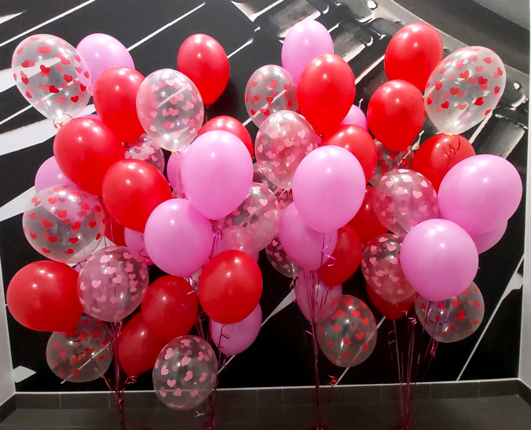 Bukiet czerwonych i różowych balonów, w tym przezroczystych z nadrukiem małych czerwonych serduszek, na tle ściany w czarno-białe pasy.