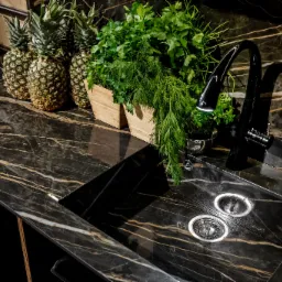 Czarny blat kuchenny z marmurowym wzorem i złotymi żyłami, z wbudowanym zlewozmywakiem i czarną baterią. Na blacie stoją trzy ananasy oraz drewniane doniczki z ziołami.