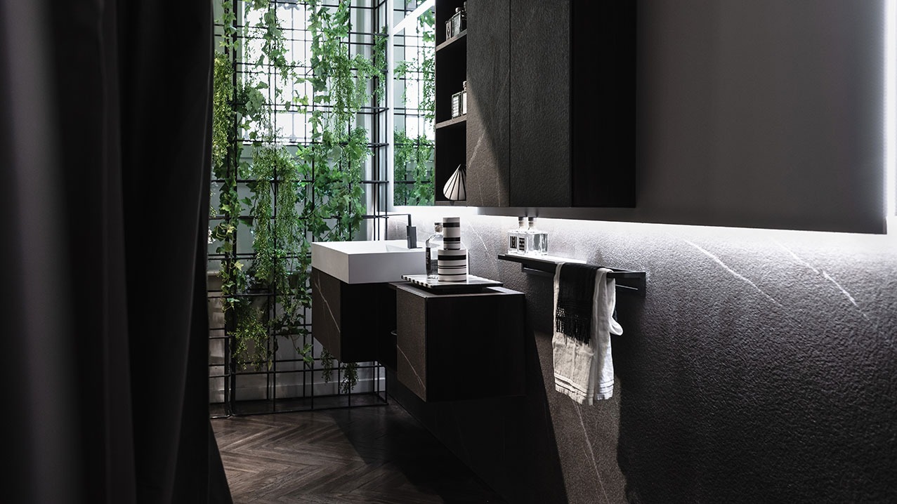 Nowoczesna łazienka z ciemnymi szafkami i blatem z efektem kamienia, białą umywalką, podświetleniem LED i ścianą z wyraźną fakturą, uzupełniona o rośliny pnące się po metalowej konstrukcji.