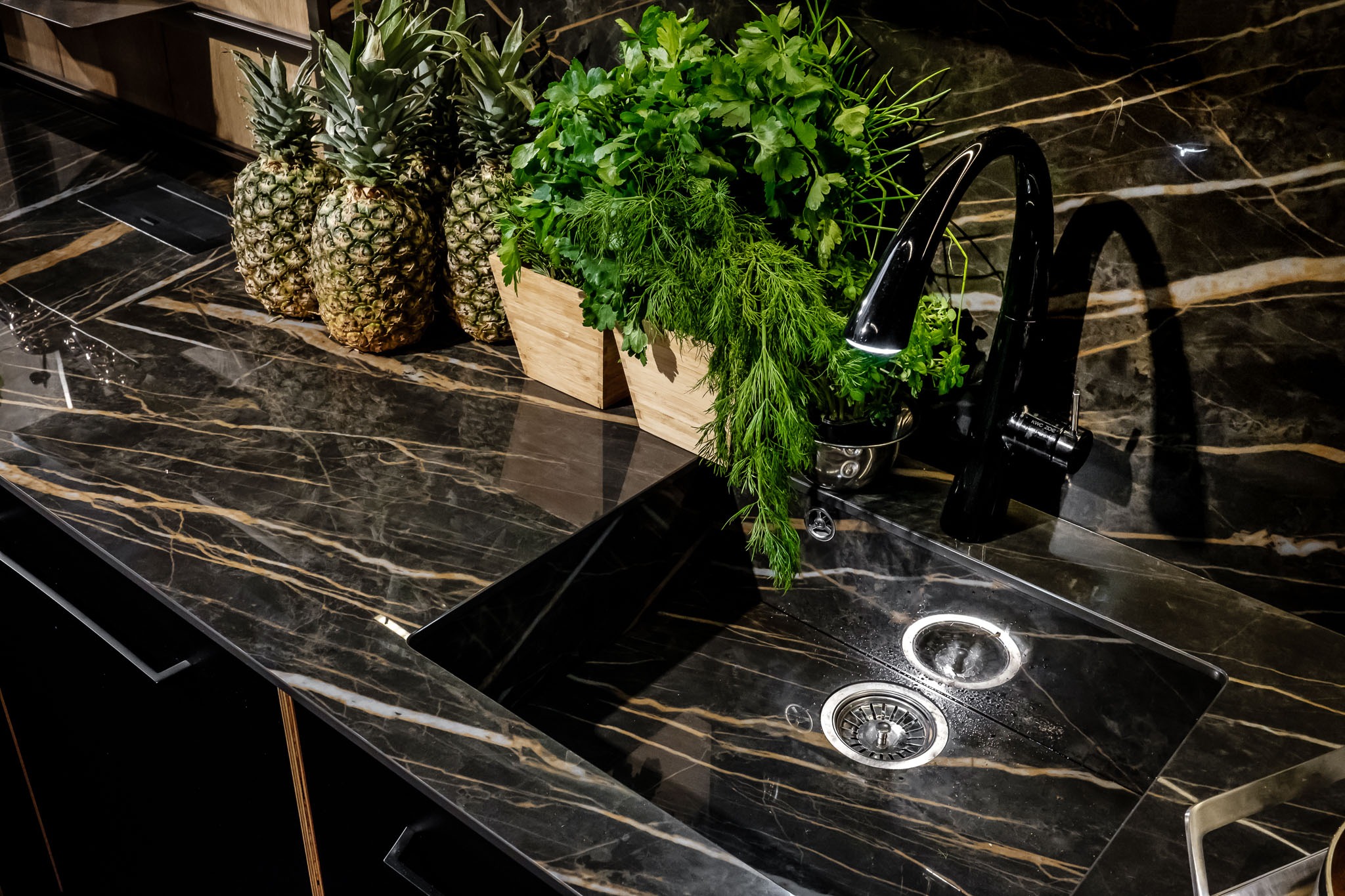 Czarny blat kuchenny z marmurowym wzorem i złotymi żyłami, z wbudowanym zlewozmywakiem i czarną baterią. Na blacie stoją trzy ananasy oraz drewniane doniczki z ziołami.