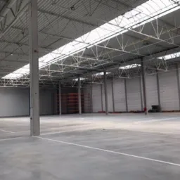 Wnętrze pustej hali magazynowej z betonową posadzką, stalową konstrukcją dachu i świetlikami, pomalowane na szaro ściany z widocznymi drzwiami po lewej stronie.