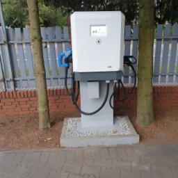 Stacja ładowania pojazdów elektrycznych marki ABB, umieszczona na betonowej podstawie z drobnym żwirem, z dwoma kablami ładującymi i wyświetlaczem, otoczona drzewami i ogrodzeniem.