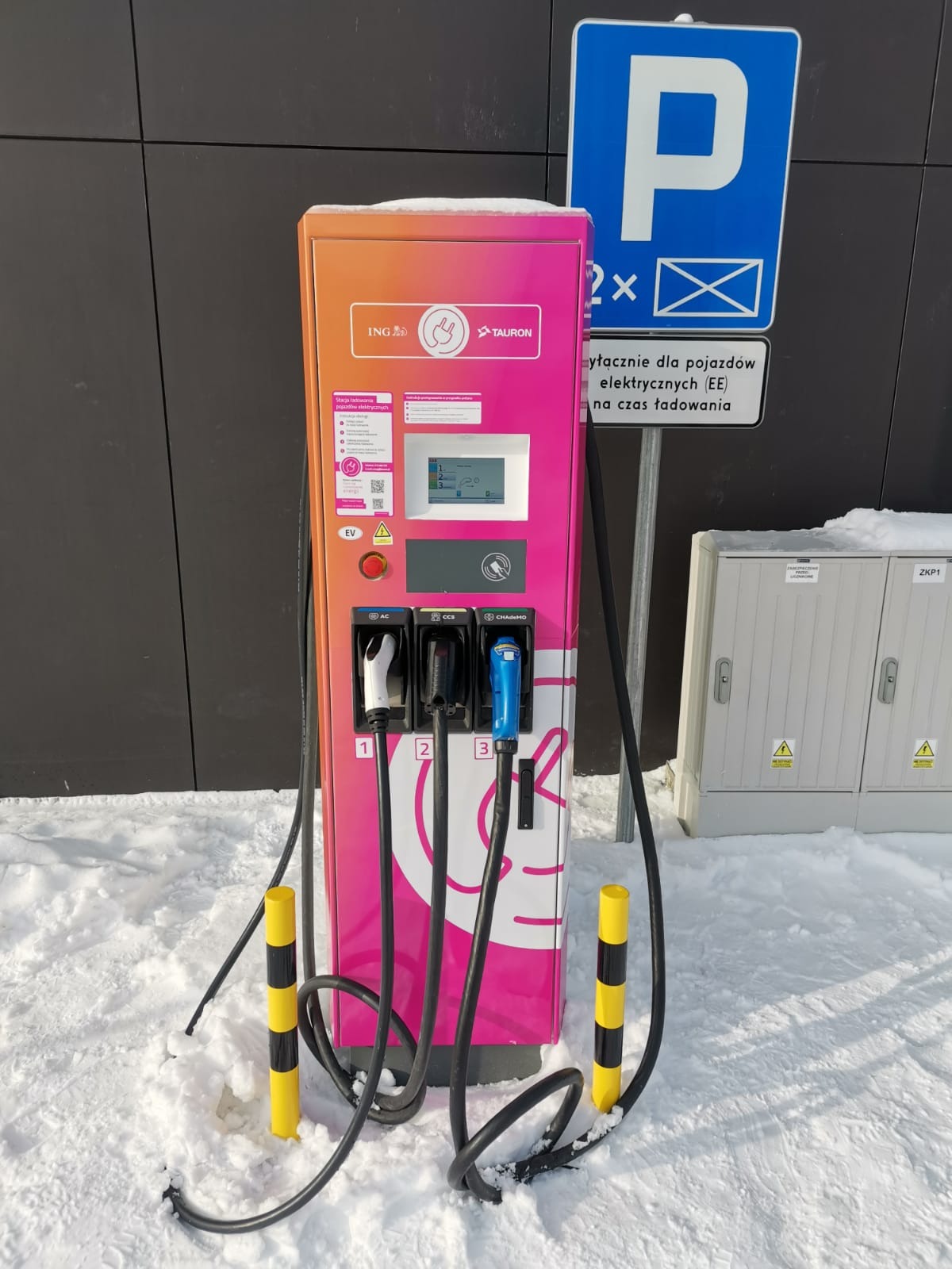 Stacja ładowania pojazdów elektrycznych z trzema złączami (AC, CCS, CHAdeMO) w kolorach pomarańczowo-różowych z logotypami ING i Tauron, umieszczona na zewnątrz w zimowej scenerii, obok znak...