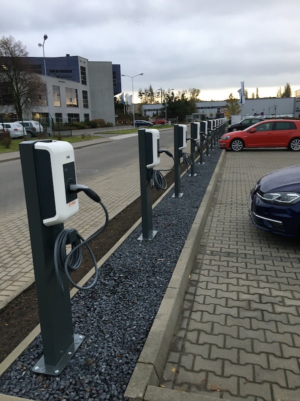 Rząd stacji ładowania pojazdów elektrycznych ABB na parkingu z kostki brukowej, widoczne zaparkowane samochody i budynek biurowy w tle, pochmurne niebo.