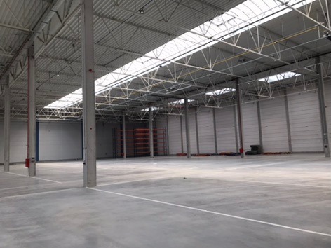 Wnętrze pustej hali magazynowej z betonową posadzką, stalową konstrukcją dachu i świetlikami, pomalowane na szaro ściany z widocznymi drzwiami po lewej stronie.