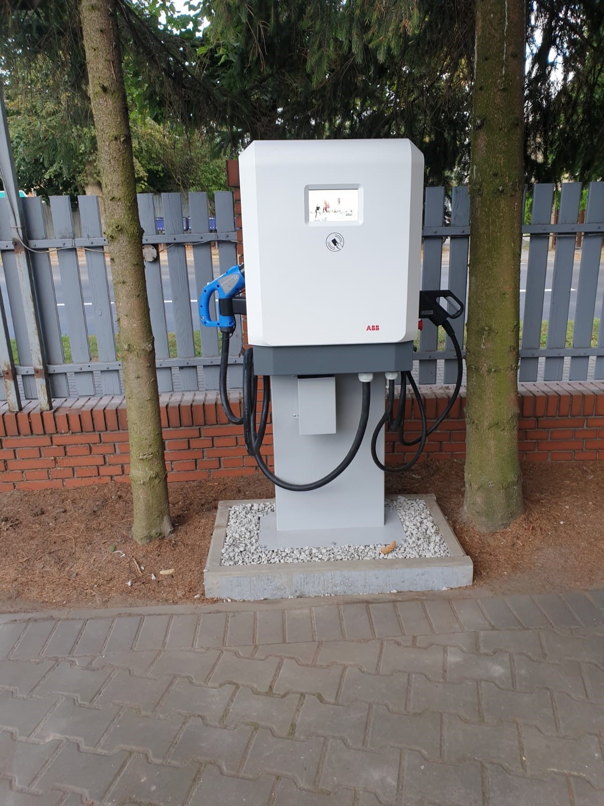 Stacja ładowania pojazdów elektrycznych marki ABB, umieszczona na betonowej podstawie z drobnym żwirem, z dwoma kablami ładującymi i wyświetlaczem, otoczona drzewami i ogrodzeniem.