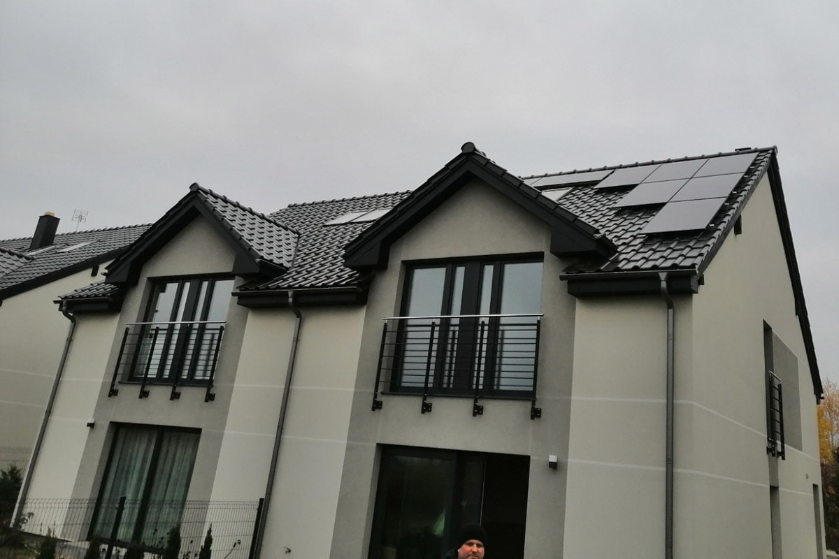 Panele słoneczne zamontowane na dachu dwupiętrowego domu z balkonami i szarą elewacją, widoczne rynny i okna dachowe.