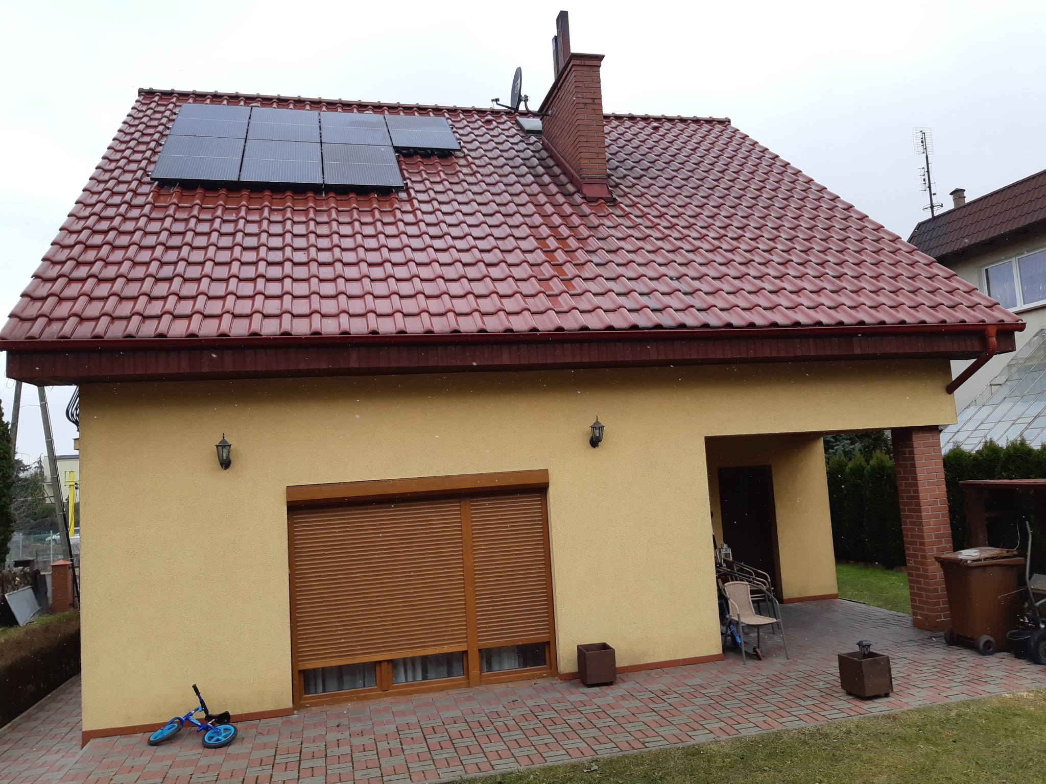 Żółty dom jednorodzinny z czerwoną dachówką, na dachu zamontowane panele fotowoltaiczne, obok komin z anteną satelitarną, na podjeździe leży niebieski rowerek biegowy.