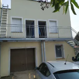 Dwupiętrowy budynek z balkonem i zewnętrznymi schodami, z drabiną opartą o balkon i zaparkowanymi samochodami.