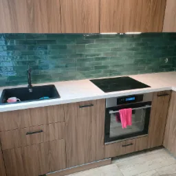 Nowoczesna kuchnia z drewnianymi szafkami, blatem w jasnym odcieniu i zielonymi, błyszczącymi płytkami na ścianie, z czarnym zlewem i baterią oraz piekarnikiem z różowym ręcznikiem na uchwycie.