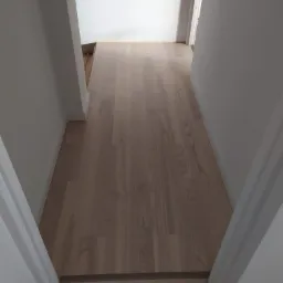 Jasne panele podłogowe ułożone w korytarzu, widoczne białe listwy przypodłogowe i ościeżnice drzwi, subtelne przejście kolorystyczne paneli przy progu.