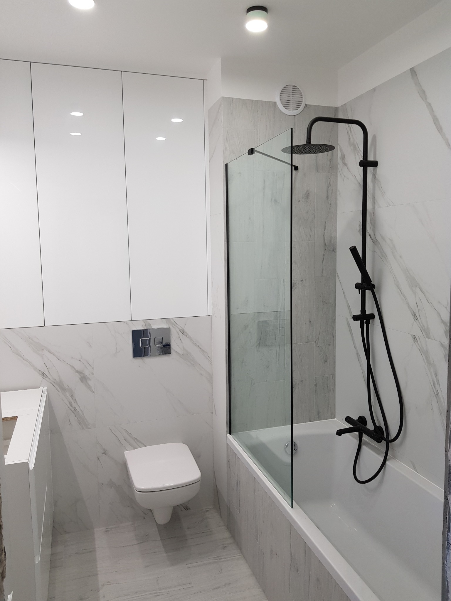 Nowoczesna łazienka z białymi szafkami, marmurowymi płytkami i czarną armaturą prysznicową za szklanym panelem.