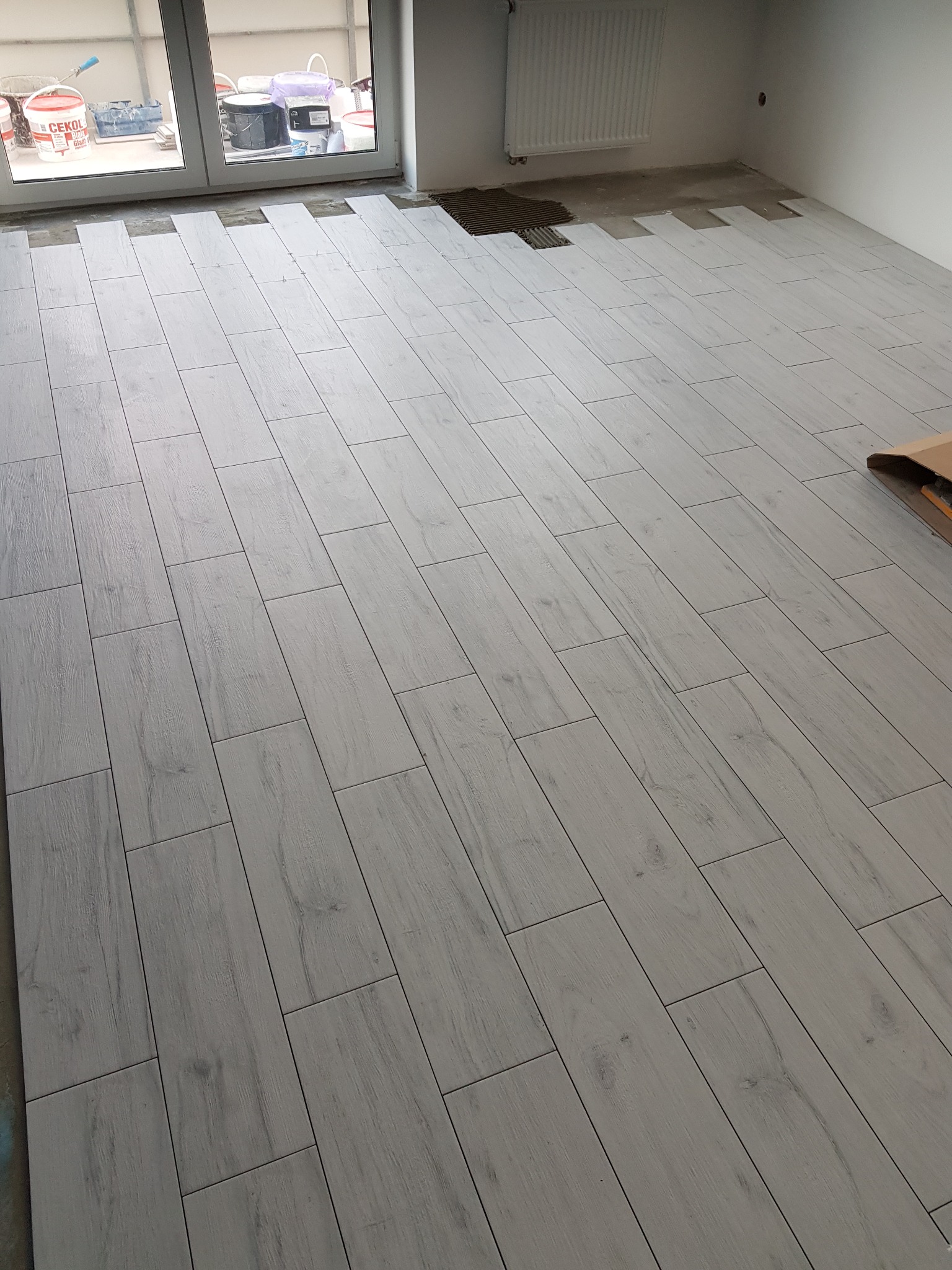 Podłoga z jasnych, podłużnych płytek imitujących drewno, ułożonych w jodełkę, z widocznymi pozostałościami kleju i workami z zaprawą w tle przy oknie.