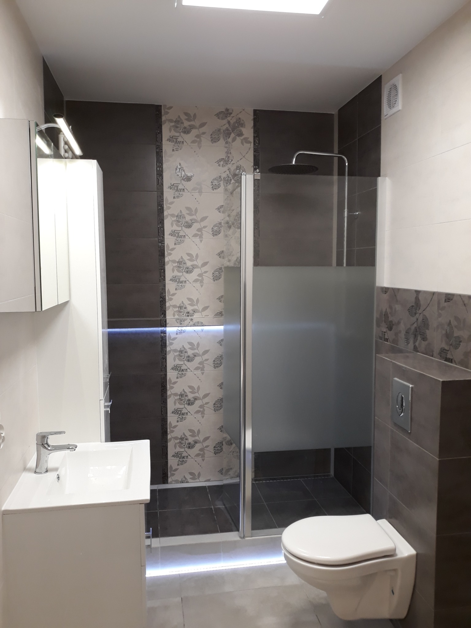 Nowoczesna łazienka z kabiną prysznicową typu walk-in z częściowo matową szybą, wiszącą toaletą, umywalką z szafką i dekoracyjnymi płytkami ściennymi z motywem liści.