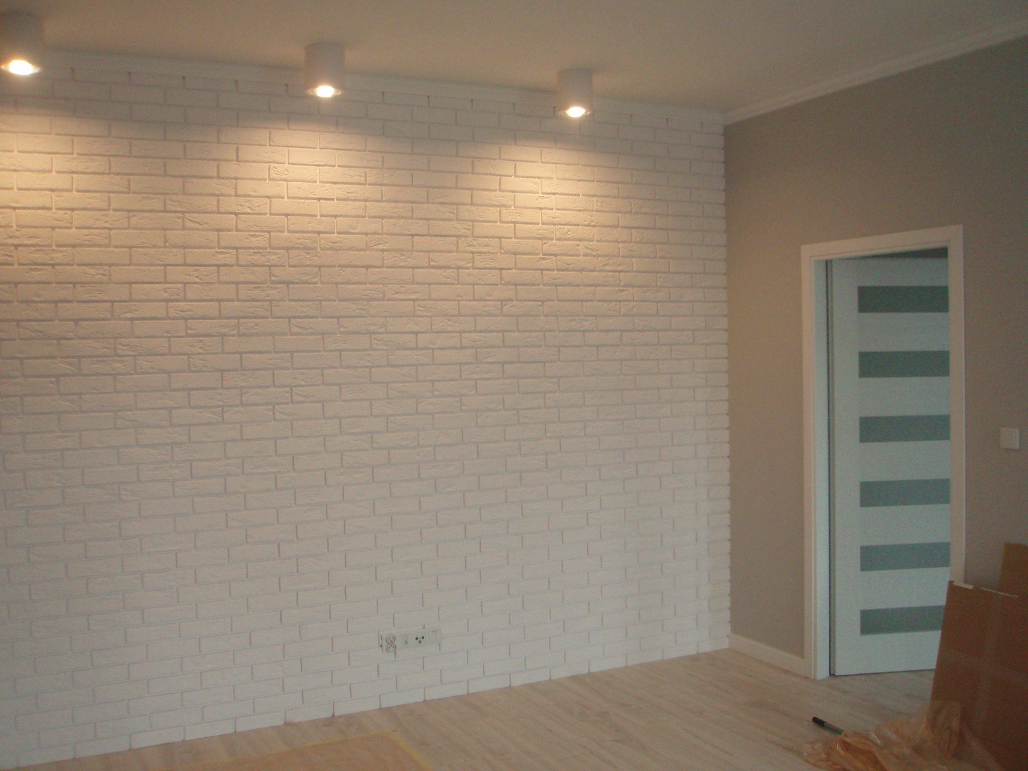 Ściana wyłożona białą cegłą dekoracyjną, oświetlona trzema punktowymi lampami sufitowymi, z widocznym gniazdkiem elektrycznym i fragmentem podłogi panelowej oraz drzwi wewnętrznych z poziomymi...