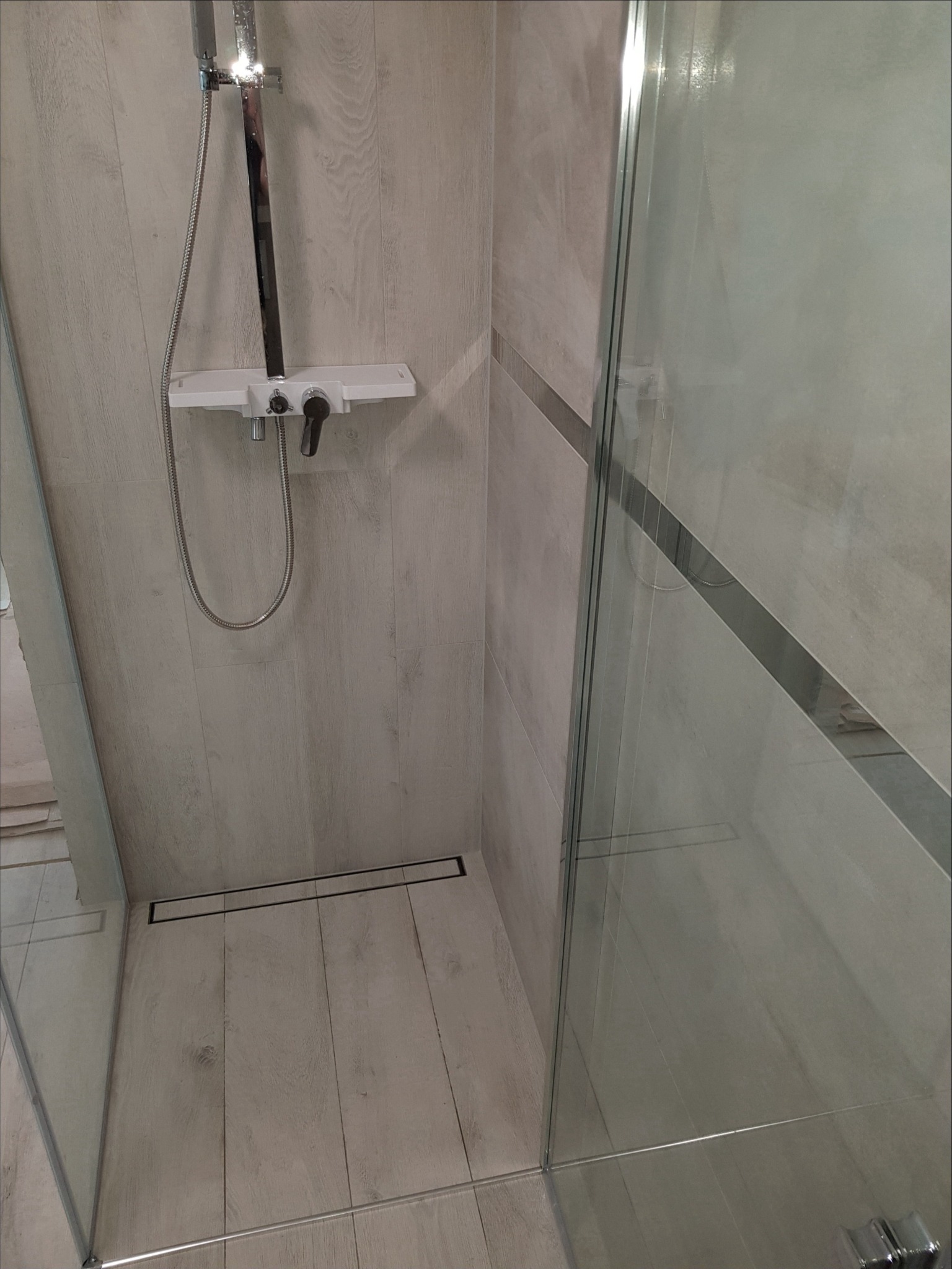 Kabina prysznicowa z odpływem liniowym w posadzce wyłożonej płytkami imitującymi drewno, widoczna bateria i deszczownica.