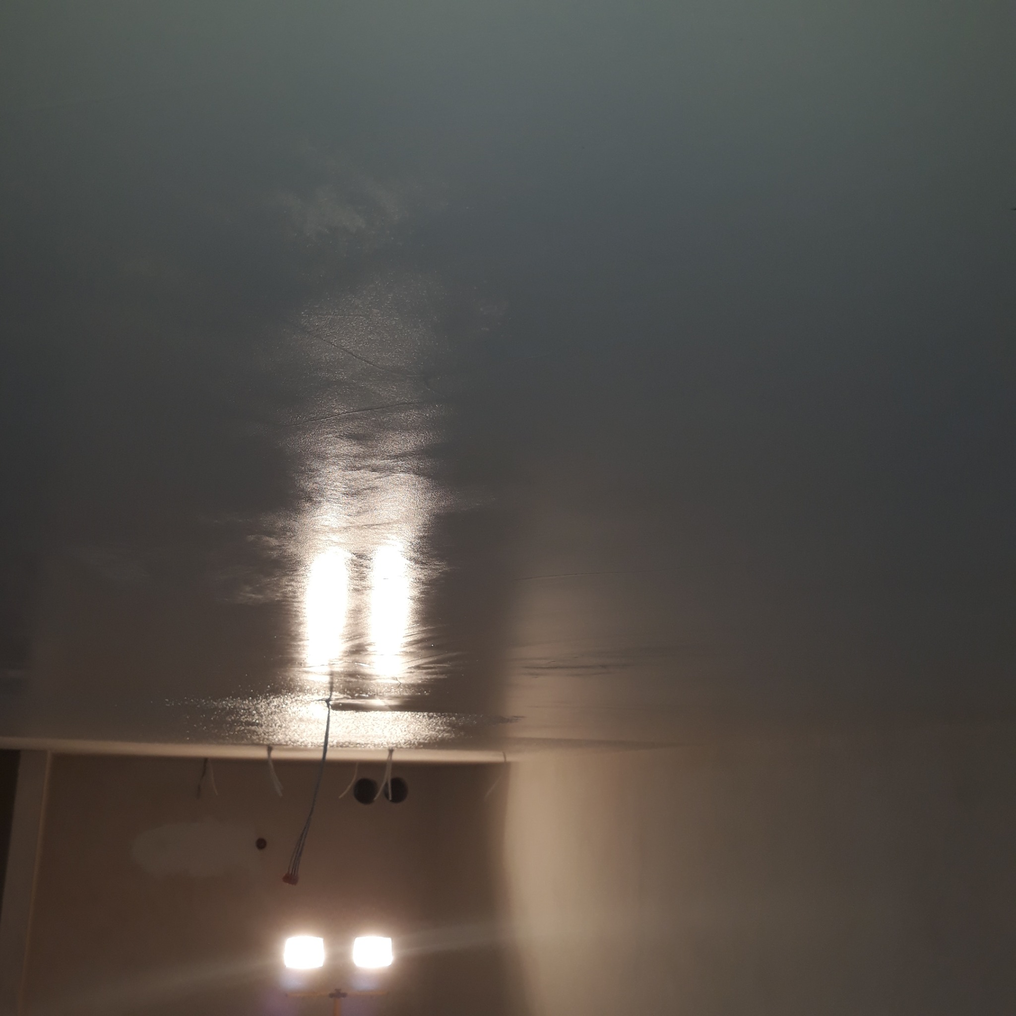 Świeżo pomalowany sufit na szaro z widocznymi smugami i odbiciami światła z lamp halogenowych. W tle widoczne wystające kable i przygotowana ściana do dalszych prac wykończeniowych.