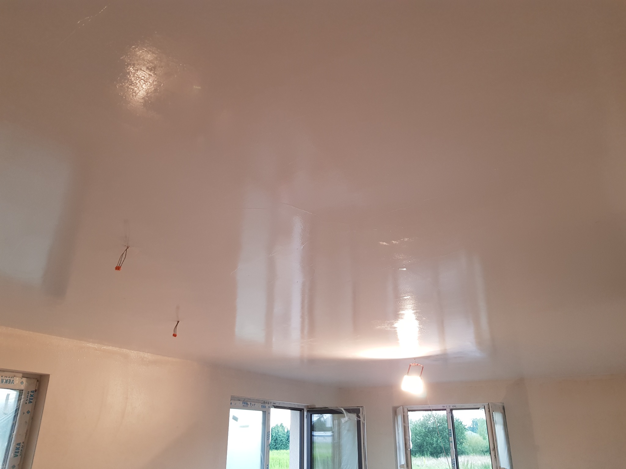 Świeżo pomalowany, lśniący sufit w jasnym odcieniu z widocznymi przewodami elektrycznymi, oświetlony lampą budowlaną, z oknami VEKA w tle.