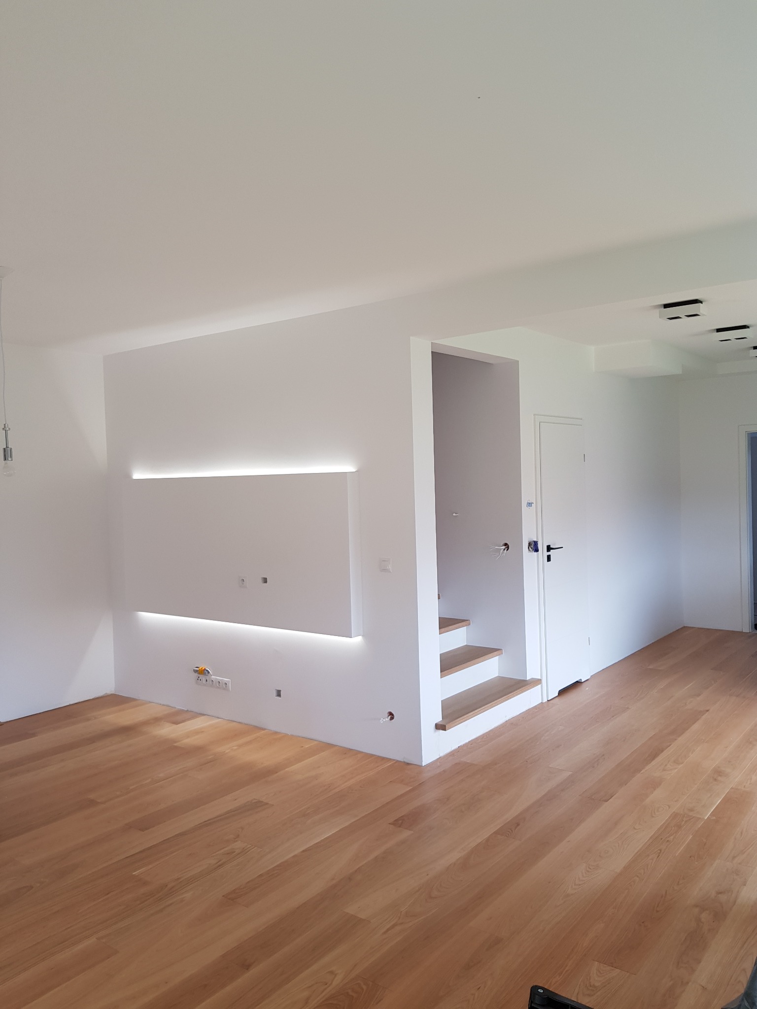 Jasne wnętrze po remoncie: drewniana podłoga, białe ściany z podświetleniem LED, schody z drewnianymi stopniami, białe drzwi z czarną klamką, gniazdka elektryczne przy podłodze.