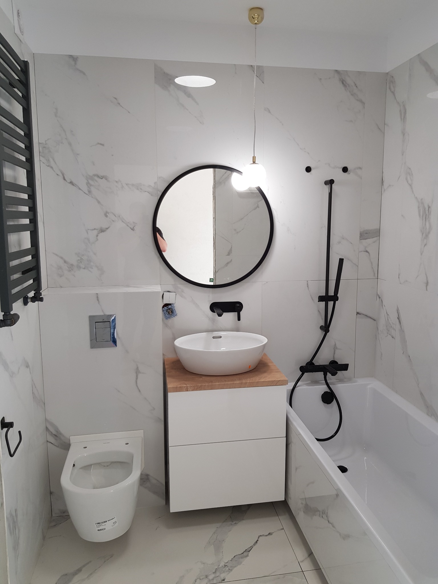 Nowoczesna łazienka z białymi płytkami imitującymi marmur, okrągłym lustrem w czarnej ramie, wiszącą toaletą i czarną armaturą.