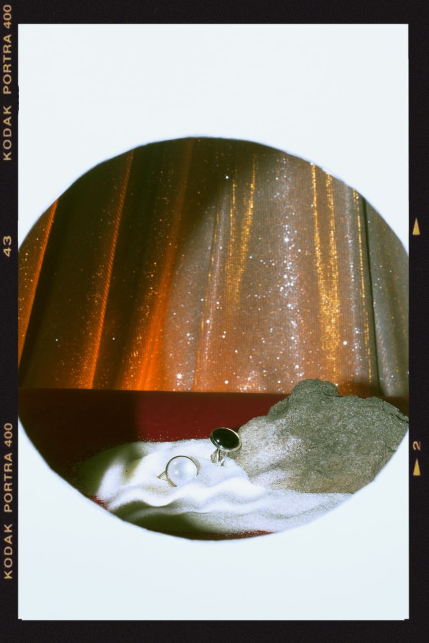 Dwa pierścionki z białym i czarnym kamieniem na piasku, obok szarego kamienia, na tle błyszczącej, pomarańczowej zasłony, ujęcie w okrągłej ramce z widocznymi oznaczeniami kliszy fotograficznej.