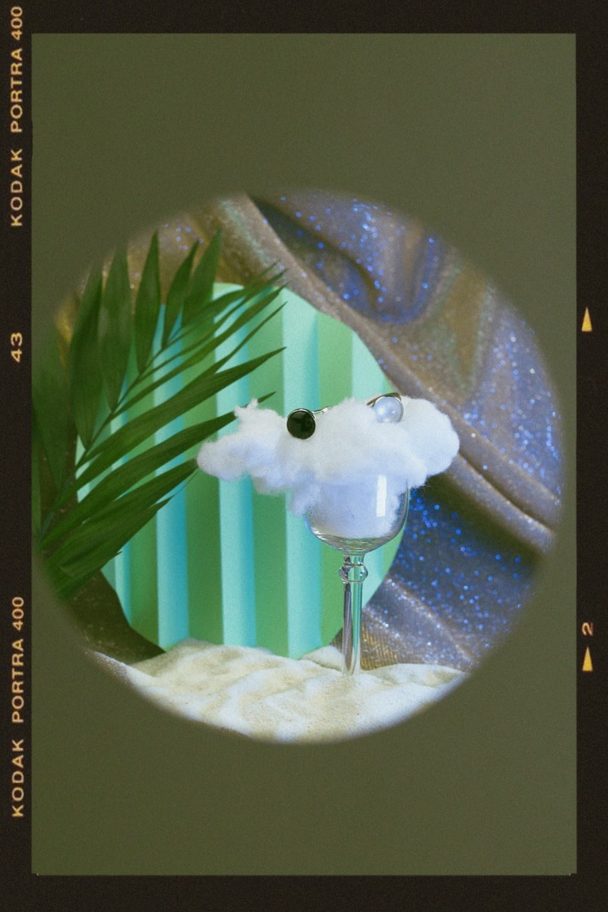 Dwa pierścionki z czarnym i białym oczkiem na chmurce z waty, umieszczonej na kieliszku, na tle zielonej palmy, błyszczącej tkaniny i zielonych pionowych pasków, w stylu vintage.