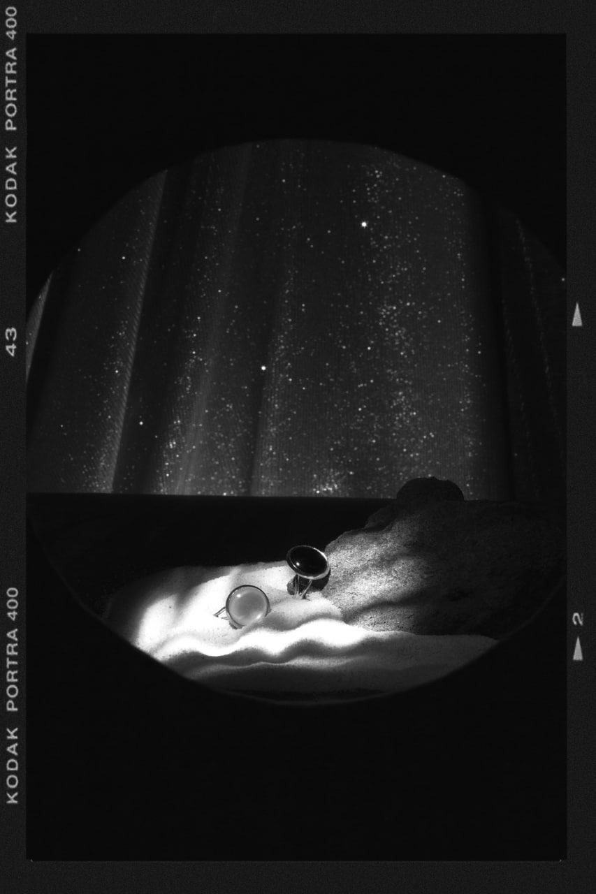 Czarno-białe zdjęcie dwóch pierścionków z kamieniami na piasku, z widocznym ziarnem kliszy i oznaczeniami Kodak Portra 400 po bokach, górna część kadru przedstawia abstrakcyjne tło przypominające...