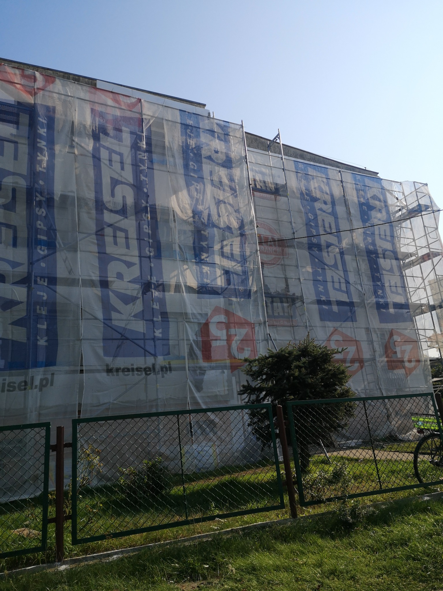 Budynek wielorodzinny w trakcie prac, osłonięty siatką z reklamą materiałów budowlanych Kreisel, widoczne rusztowania i fragment ogrodzenia z zielenią.