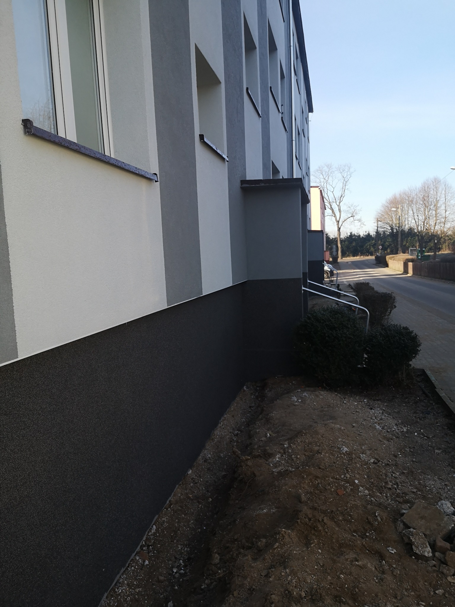 Ocieplona elewacja budynku wielorodzinnego z wyraźnym podziałem na część dolną w ciemnym kolorze i górną w jasnym odcieniu szarości, widoczne metalowe parapety i fragment chodnika z ziemią.
