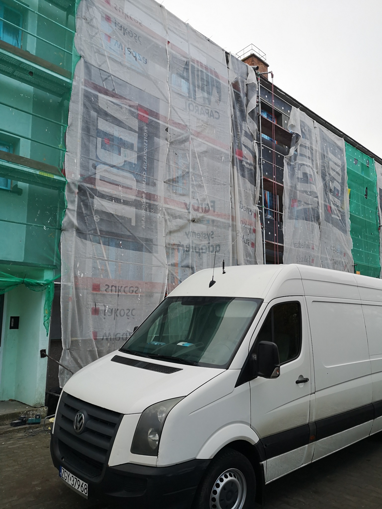 Budynek w trakcie prac elewacyjnych, osłonięty siatką rusztowaniową z widocznym fragmentem ocieplenia i zaparkowanym białym vanem firmy.