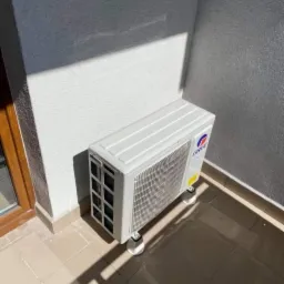 Kompaktowa jednostka zewnętrzna klimatyzacji marki Gree, umieszczona na niskich nóżkach, stoi na wyłożonym płytkami balkonie, obok okna, z widocznym cieniem rzucanym przez słońce na ścianę.