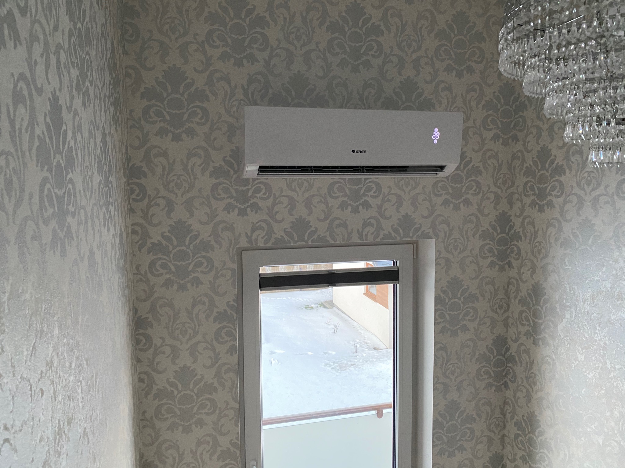 Biały klimatyzator marki Gree zamontowany na ścianie w pokoju z tapetą w szaro-białe ornamenty, widoczny kryształowy żyrandol i okno z zimowym widokiem.