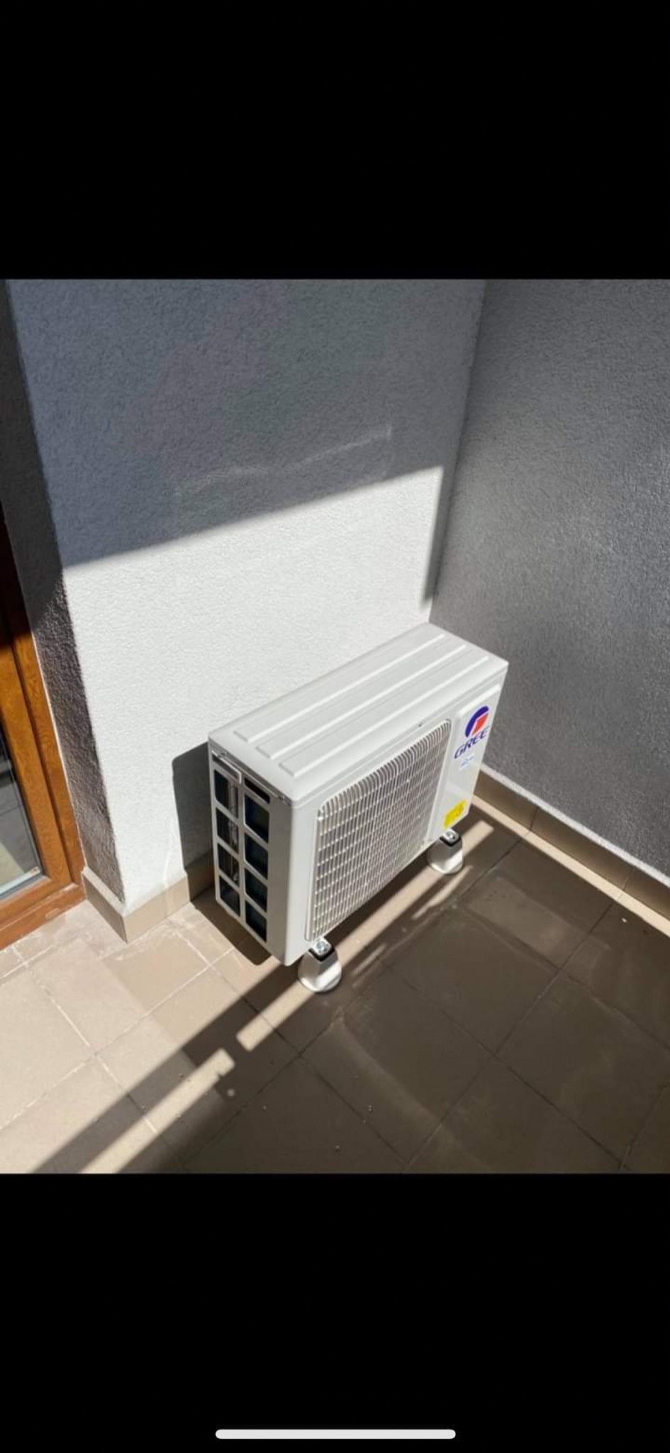 Kompaktowa jednostka zewnętrzna klimatyzacji marki Gree, umieszczona na niskich nóżkach, stoi na wyłożonym płytkami balkonie, obok okna, z widocznym cieniem rzucanym przez słońce na ścianę.